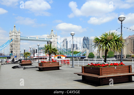 Butlers Wharf Thames Path Promenade accanto al fiume Tamigi viste del Tower Bridge e della città di Londra skyline fila alberi cordyline fiori rossi in piantatrici UK Foto Stock