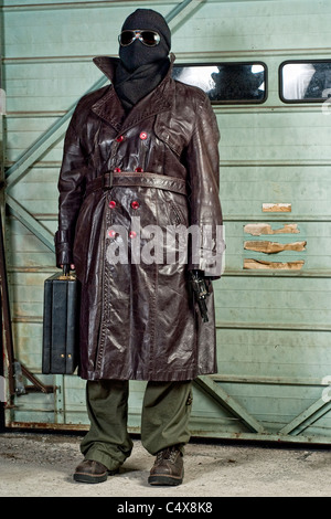 Una persona che indossa gli occhiali da sole, rivestimento in pelle e una ski-mask detiene una valigetta e una pistola. Foto Stock