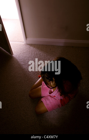 Giovane donna seduta da sola in una stanza buia indossando un rosa vestaglia guardando verso il basso. Foto Stock