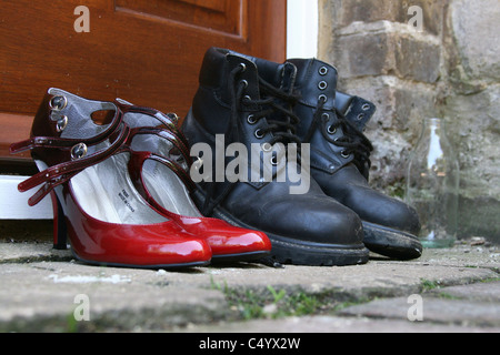 Una coppia di nero stivali da lavoro esterno su una porta accanto a una coppia di colore rosso in pelle di brevetto ladies vestono scarpe. Foto Stock