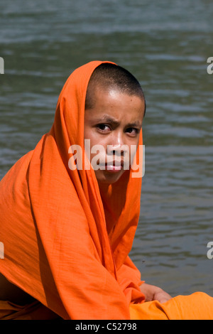 Un giovane monaco buddista è seduto in una barca con il suo manto drappeggiato sopra la sua testa sulle rive di un fiume in Laos comunista. Foto Stock