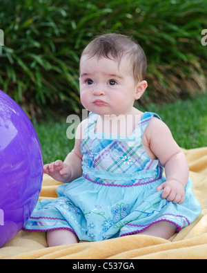 Un otto mesi baby caucasica ragazza si siede su una coperta accanto a una grande sfera di colore viola. Foto Stock
