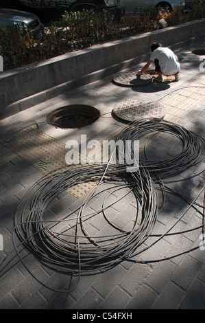 Un lavoratore cinese squat accanto a un tombino aperto / tombino e cavo / fili durante l'esecuzione di lavori di manutenzione e di riparazione. Foto Stock