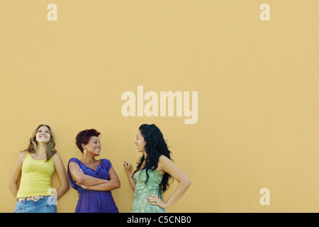 Un gruppo di tre donne amici ispanica appoggiata sulla parete gialla e ridere. Sagoma orizzontale, vita, spazio di copia Foto Stock
