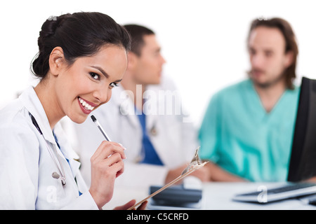 Giovani asiatici medico sorridente con la penna e guardando la telecamera, altri sfocato dietro su sfondo bianco Foto Stock