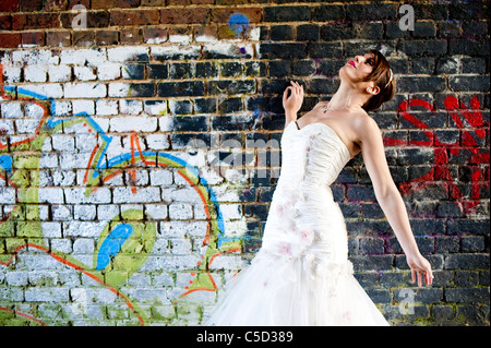 Sposa pone contro il muro di graffiti in abito da sposa guardando lontano Foto Stock