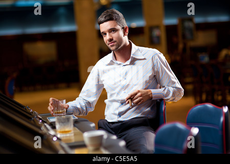 Giovani uomini seduti in casino slot machine, tenendo un sigaro cubano e un bicchiere di alcool Foto Stock