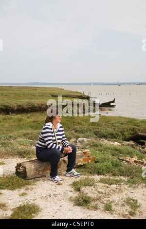 Una donna si siede su un log guardando le Swale, sull'Isle of Sheppey, Kent. Foto Stock