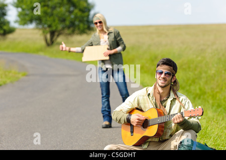 Sollevatore-escursione coppia giovane zaino tramping su strada asfaltata suonare la chitarra Foto Stock