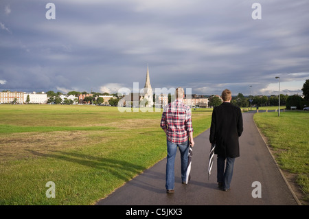 Due giovani che portano ombrelloni che attraversano la brughiera fino al villaggio la sera. Blackheath, Londra, Inghilterra, Regno Unito, Gran Bretagna Foto Stock