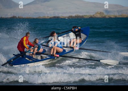Surf rescue barca a remi andando fuori sopra le onde che si infrangono con quattro donne di canottaggio e un uomo sul timone, Lyall Bay, Wellington, Nuova Foto Stock