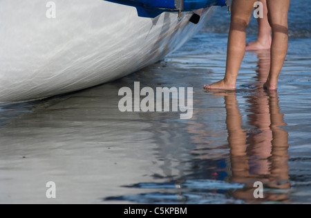 Gambe e piedi di due donne accanto un surf rescue barca a remi sulla sabbia bagnata, Lyall Bay, Wellington, Nuova Zelanda Foto Stock