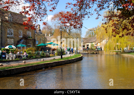 I turisti in sole primaverile accanto al Fiume Windrush, Bourton sull'acqua, Gloucestershire, Cotswolds, England, Regno Unito Foto Stock