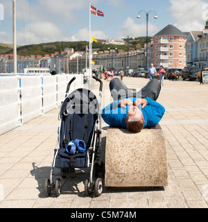 Un uomo sdraiato su un banco di mare accanto a un vuoto kids buggy, aberystwyth wales uk Foto Stock