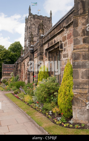 Chiesa della Santissima Trinità, Skipton (portico sud, torre, bandiera, percorso in pietra, piante e arbusti nel giardino di chiesa confine erbaceo) - Yorkshire, Inghilterra, Regno Unito. Foto Stock