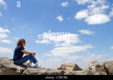 Meditando sessanta anni di donna matura si siede su rocce e sguardi fuori in un nuvoloso cielo blu. Foto Stock