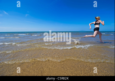 Womans jogging lungo una spiaggia sabbiosa schizzi attraverso l'Oceano surf. Foto Stock