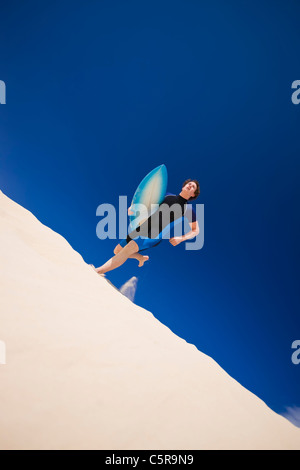 Giovane uomo che corre attraverso le dune di surf in oceano con la tavola da surf. Foto Stock