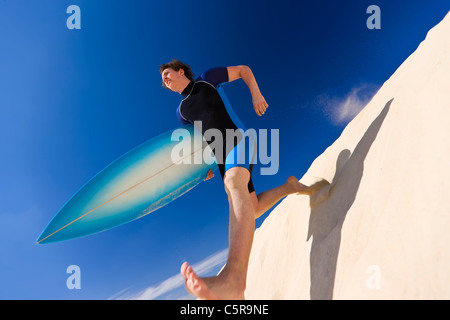 Surfer facendo grandi passi verso il basso dune di sabbia all'oceano. Foto Stock