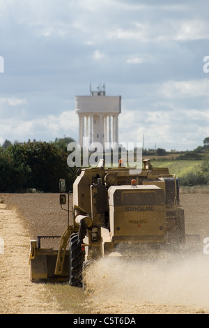 New Holland TX65 Mietitrebbia la mietitura del frumento in North Norfolk con la torre dell'acqua a Hill sessanta nella distanza. Foto Stock