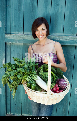 In Germania, in Baviera, donna di fronte a fienile porta cestello di contenimento con verdure fresche, sorridente, ritratto Foto Stock