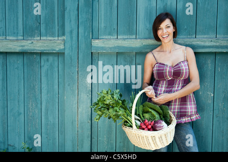 In Germania, in Baviera, donna in piedi di fronte a porta del granaio, cestello di contenimento con verdure fresche, sorridente, ritratto Foto Stock