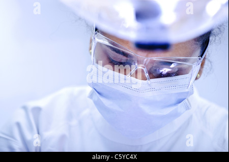 In prossimità della faccia del giovane femmina dentista indossare una mascherina chirurgica e occhiali e bianco tunica dentale guardando verso il basso Foto Stock