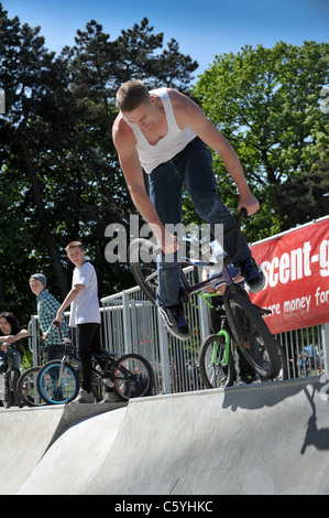 Maschio bianco gioventù in un corpetto su uno stunt bike salta fuori da una rampa a skate park guardato da altri ciclisti adolescente Foto Stock