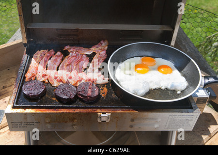 Uova fritte e pancetta e black pudding cottura su un portatile barbecue a gas. Foto Stock