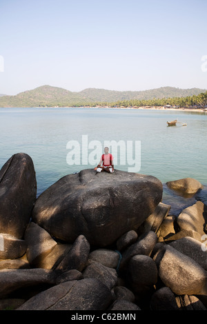 Un uomo indiano le pratiche yoga meditazione su una roccia presso la popolare località turistica di Palolem Beach nel sud dello stato di Goa, India Foto Stock