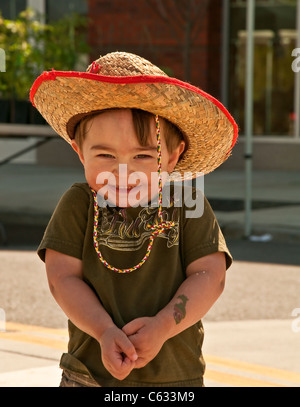 Grazioso, piccolo felice 2 anno vecchio toddler boy, sorridente con le sue spalle scrollato le spalle. Egli indossa un vecchio paglia cappello da cowboy. Foto Stock