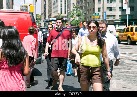 Folla variegata pedoni vestito per una calda giornata estiva cross 57th street presso la Sesta Avenue nel centro di Manhattan a New York City Foto Stock