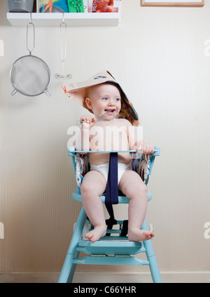 6 mesi di età bambino seduto nella sedia bambino indossando un cappello di giornale Foto Stock