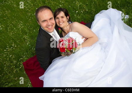 Classical coppia di novelli sposi con abito da sposa abito scuro e rosso bouquet nuziale lo sposo e la sposa giacciono su un prato Foto Stock