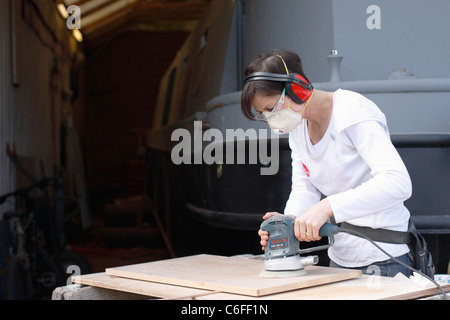 Una donna la levigatura di una porta di legno a tralicci, indossando le cuffie di protezione, occhiali di sicurezza, guanti in lattice e una maschera facciale per la protezione. Foto Stock