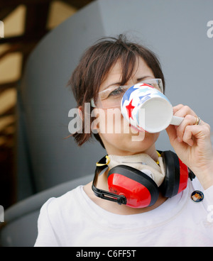 Una donna di bere una tazza di tè mentre avente una pausa dal lavoro. Indossare protezioni per orecchie e una maschera facciale per la sicurezza. Foto Stock