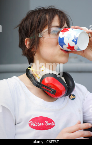 Una donna di bere una tazza di tè mentre avente una pausa dal lavoro. Indossare protezioni per orecchie e una maschera facciale per la sicurezza. Foto Stock