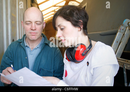 Un marito e una moglie guardando sopra alcune pratiche per il loro business, davanti a scafo in acciaio barca stretta in magazzino. Foto Stock