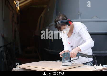 Una donna la levigatura di una porta di legno a tralicci, indossando le cuffie di protezione, occhiali di sicurezza, guanti in lattice e una maschera facciale per la protezione. Foto Stock