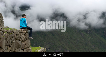Donna seduta su un muro di pietra e guardando a valle presso la cittadella Inca di Machu Picchu, regione di Cusco, Perù Foto Stock