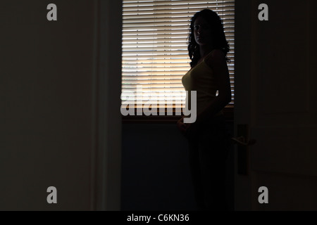 Silhouette di una giovane donna in piedi in una stanza buia la luce da una bindow dietro di lei. Foto Stock