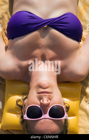 La donna a prendere il sole in bikini viola e rosa occhiali da sole, giacente su giallo asciugamano. Foto Stock