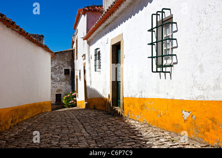 Stretta viuzza acciottolata nel vecchio centro medievale Obidos village, Portogallo. Case con pareti bianche e strisce gialle. Foto Stock