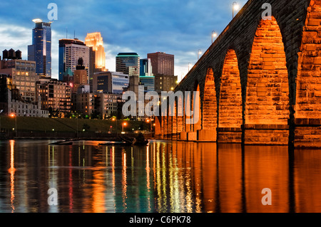 Lo skyline di Minneapolis e arco in pietra bridge al tramonto con il fiume Mississippi in primo piano. Foto Stock