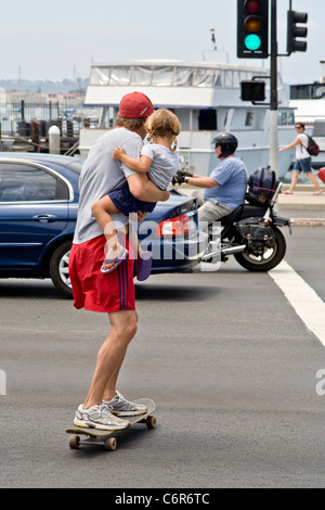 Un padre porta il suo bimbo figlia durante l'attraversamento di una strada su uno skateboard a San Diego, CA. Foto Stock