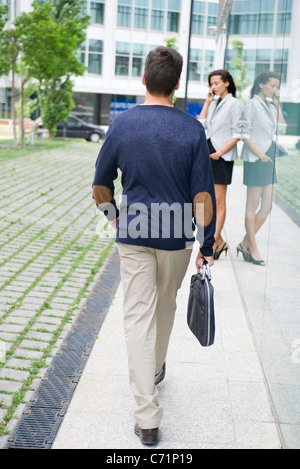 Uomo che cammina con valigetta, donna parlando al cellulare in background Foto Stock