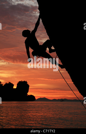 Silhouette di rocciatore salendo una rupe a strapiombo con tramonto sull'oceano sullo sfondo Foto Stock