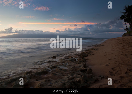 Un bellissimo tramonto in Maui con nuvole in lontananza e onde che lambiscono sui coralli sulla spiaggia Foto Stock