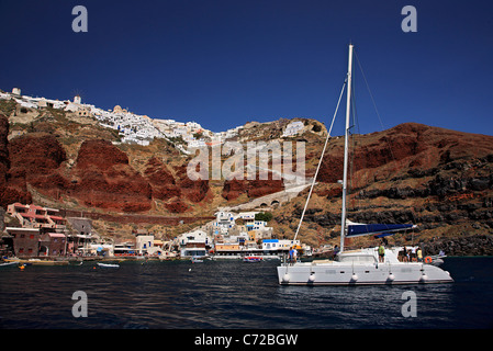 Uno yacht (catamarano) passando davanti al villaggio di Oia e Ammoudi, uno dei suoi due porti, isola di Santorini, Cicladi Grecia Foto Stock