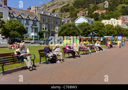 I cittadini anziani occupano panche per godersi il sole e le viste sul lungomare di Llandudno, in background è una gita in treno su strada Foto Stock